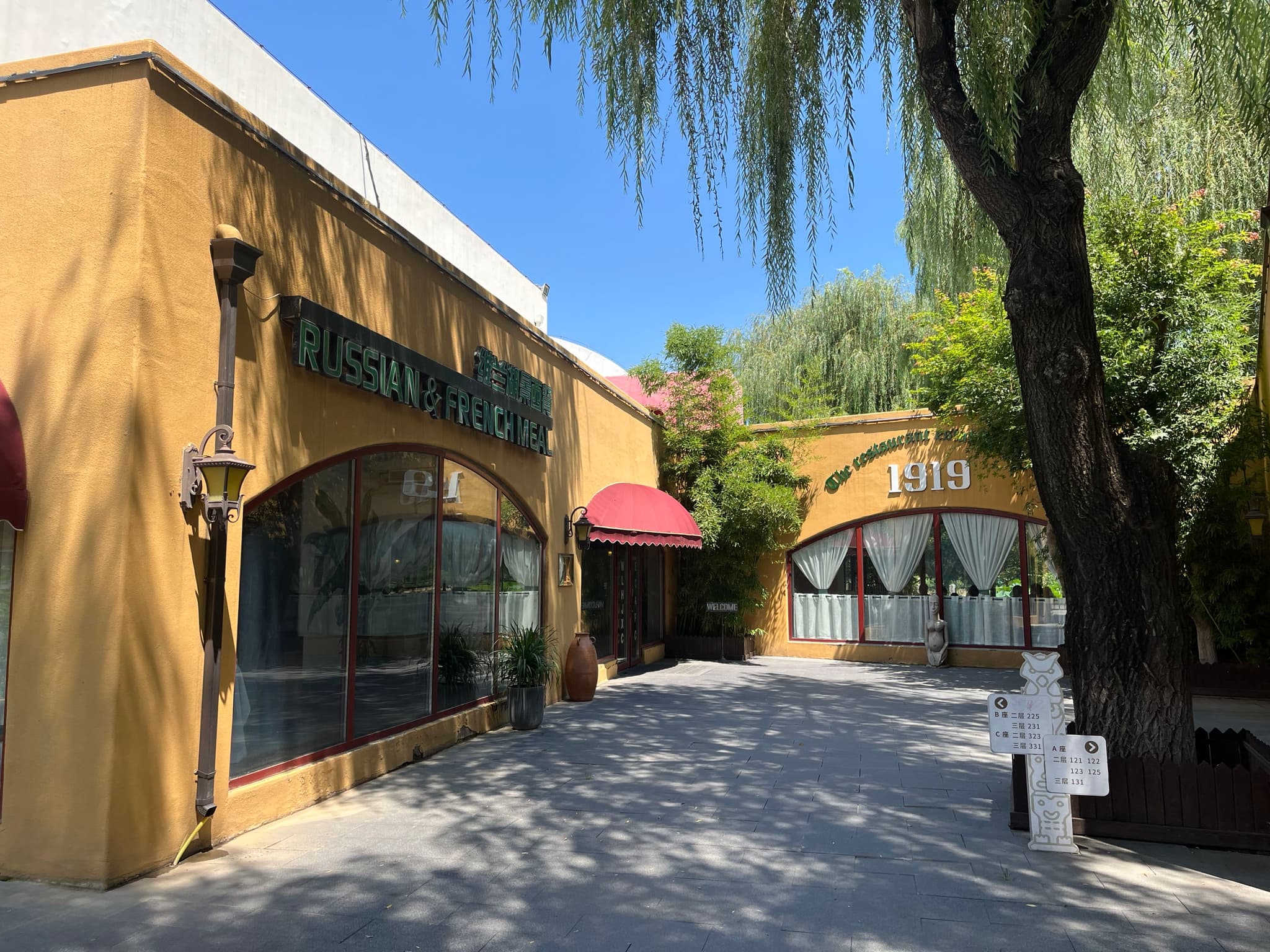 A rustic yellow building with arched windows, a red awning, and surrounding greenery under a clear blue sky