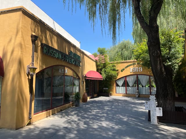 A rustic yellow building with arched windows, a red awning, and surrounding greenery under a clear blue sky