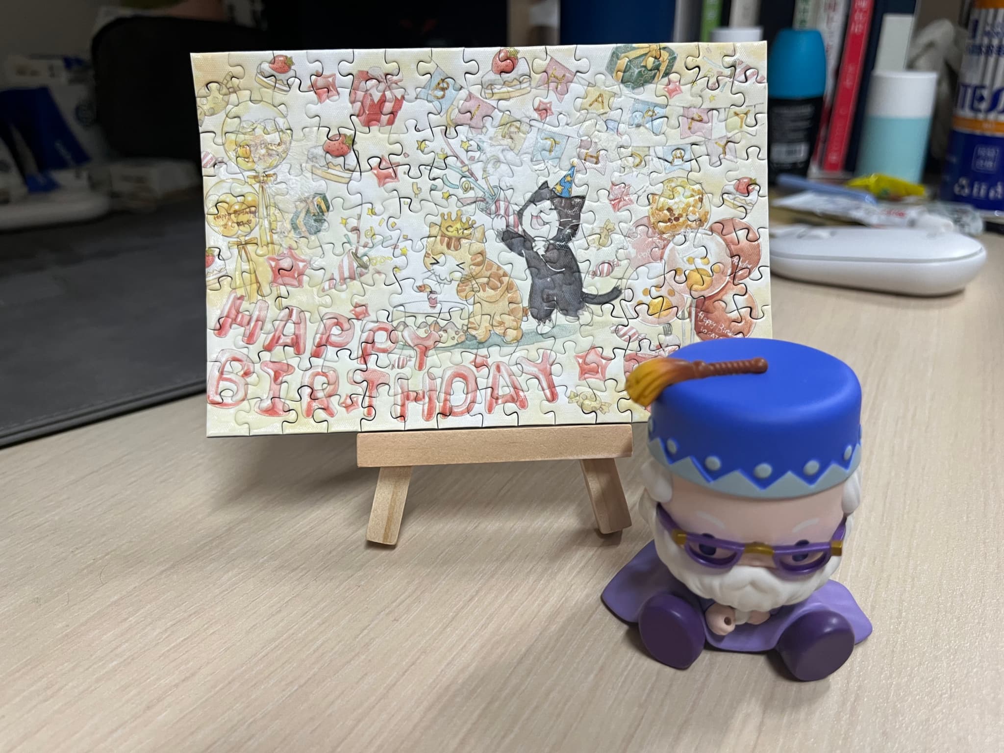 A small birthday card featuring a black cat on an easel, accompanied by a purple figurine with a blue crown and glasses on a desk