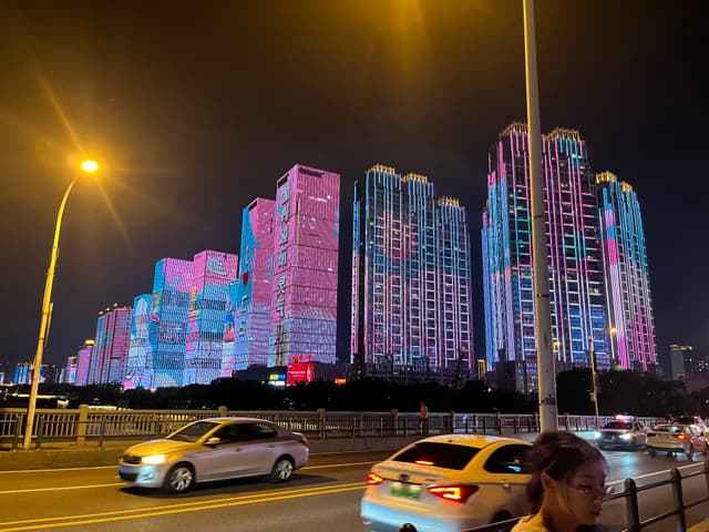 High-rise buildings illuminated with vibrant pink and blue lights at night, with cars and streetlights in the foreground