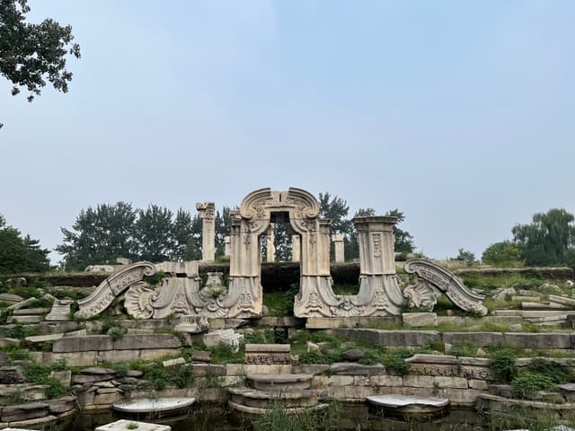 Ornate stone ruins of a palace structure with overgrown vegetation and a calm body of water in the foreground