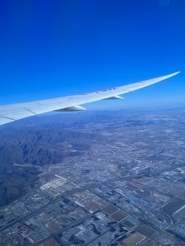 An airplane wing in flight with a view of a cityscape and fields below under a clear blue sky