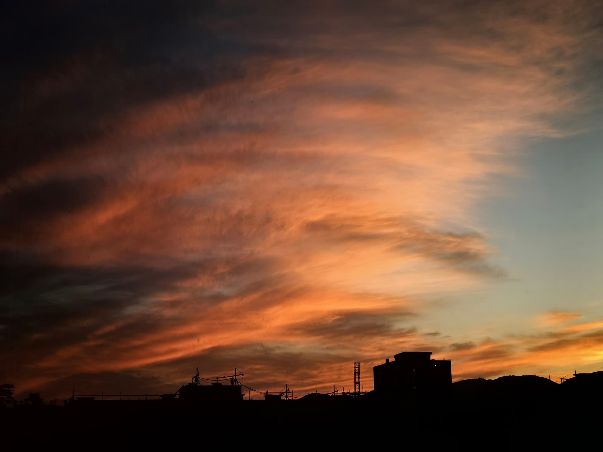 A vibrant sunset with orange and pink hues streaking across the sky, silhouetting buildings and structures on the horizon