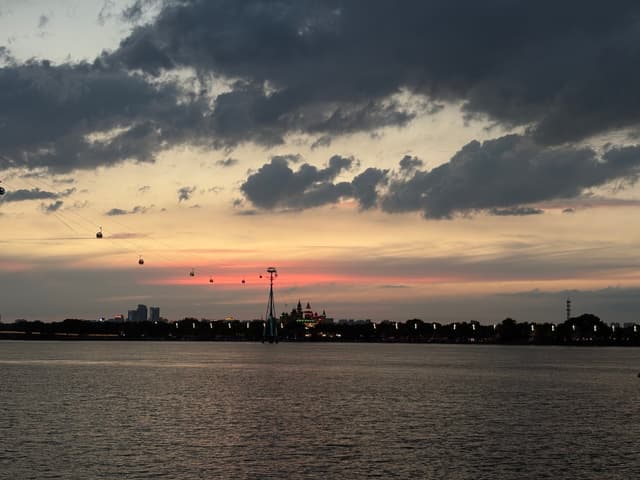 A serene waterfront at sunset with a cloudy sky, a faint pink horizon, and a line of lights along the distant shore