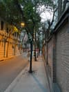 A quiet urban street lined with trees, brick walls, and dim streetlights during dusk