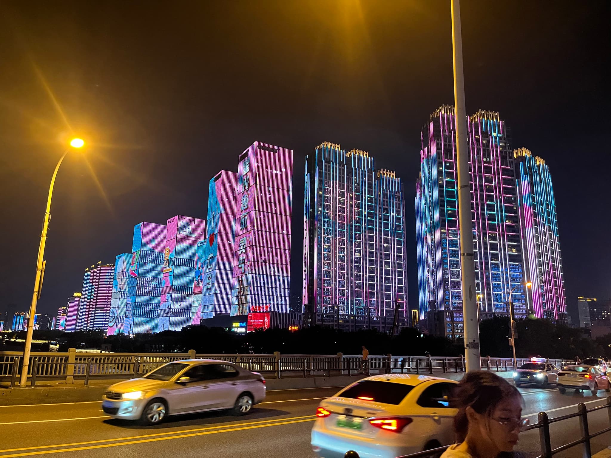 High-rise buildings illuminated with vibrant pink and blue lights at night, with cars and streetlights in the foreground