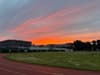 A vibrant sunrise paints the sky with orange and pink hues over a grassy field and buildings
