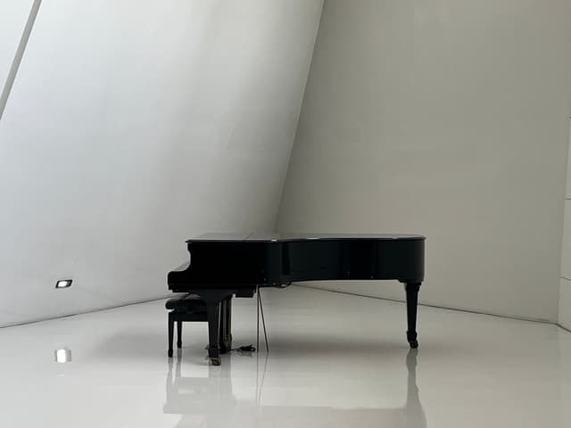A black grand piano sits on a white floor with a white wall behind it