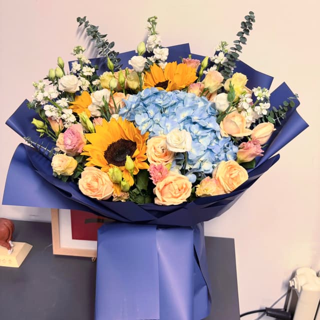A vibrant bouquet featuring sunflowers, blue hydrangeas, peach roses, and white blossoms, wrapped in deep blue paper