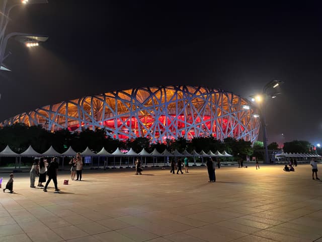 An upside-down view of a brightly lit structure with intricate lattice design, surrounded by trees and a reflective surface, under a dark sky
