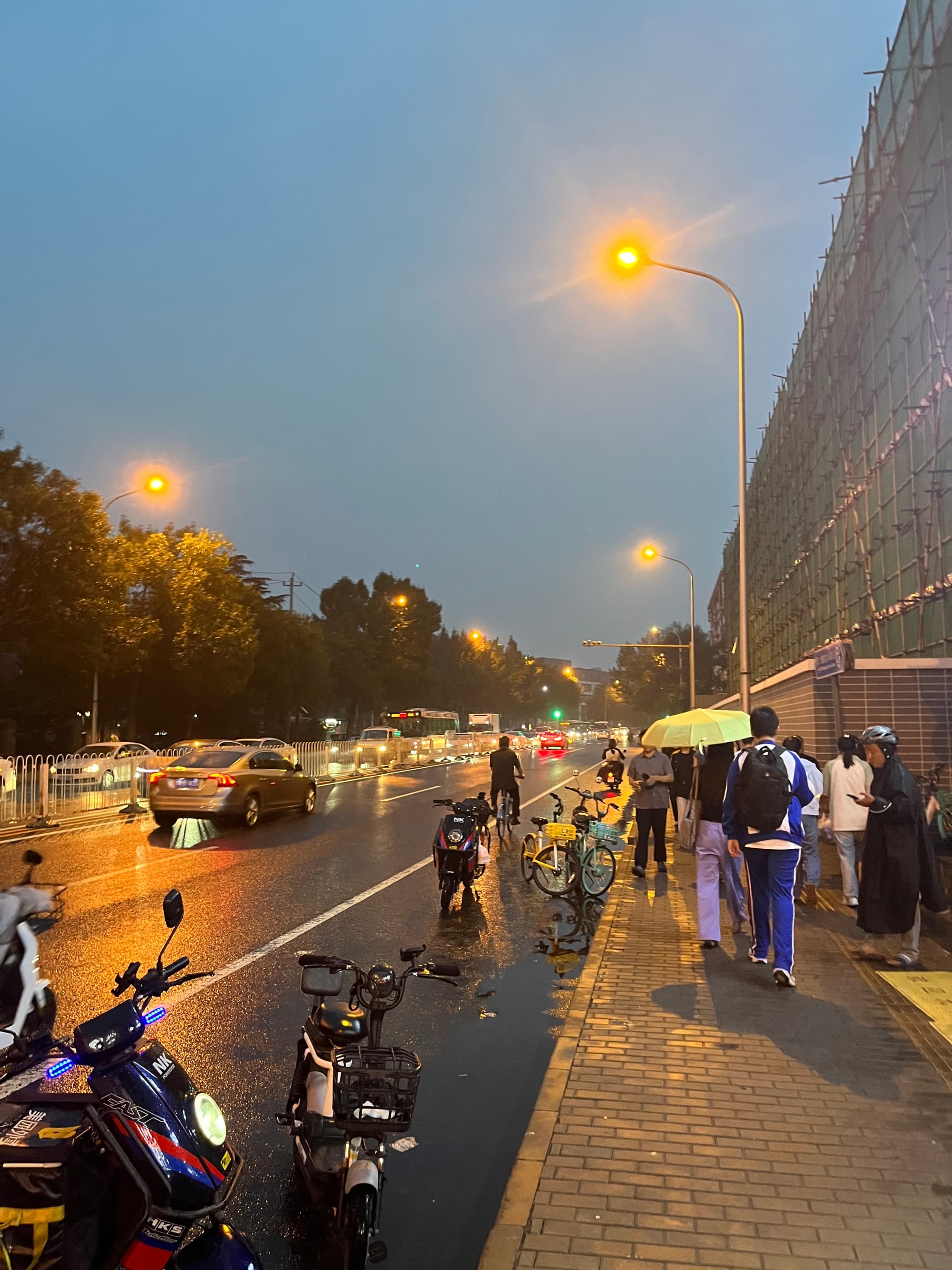 A wet street at dusk with cars and pedestrians walking under streetlights