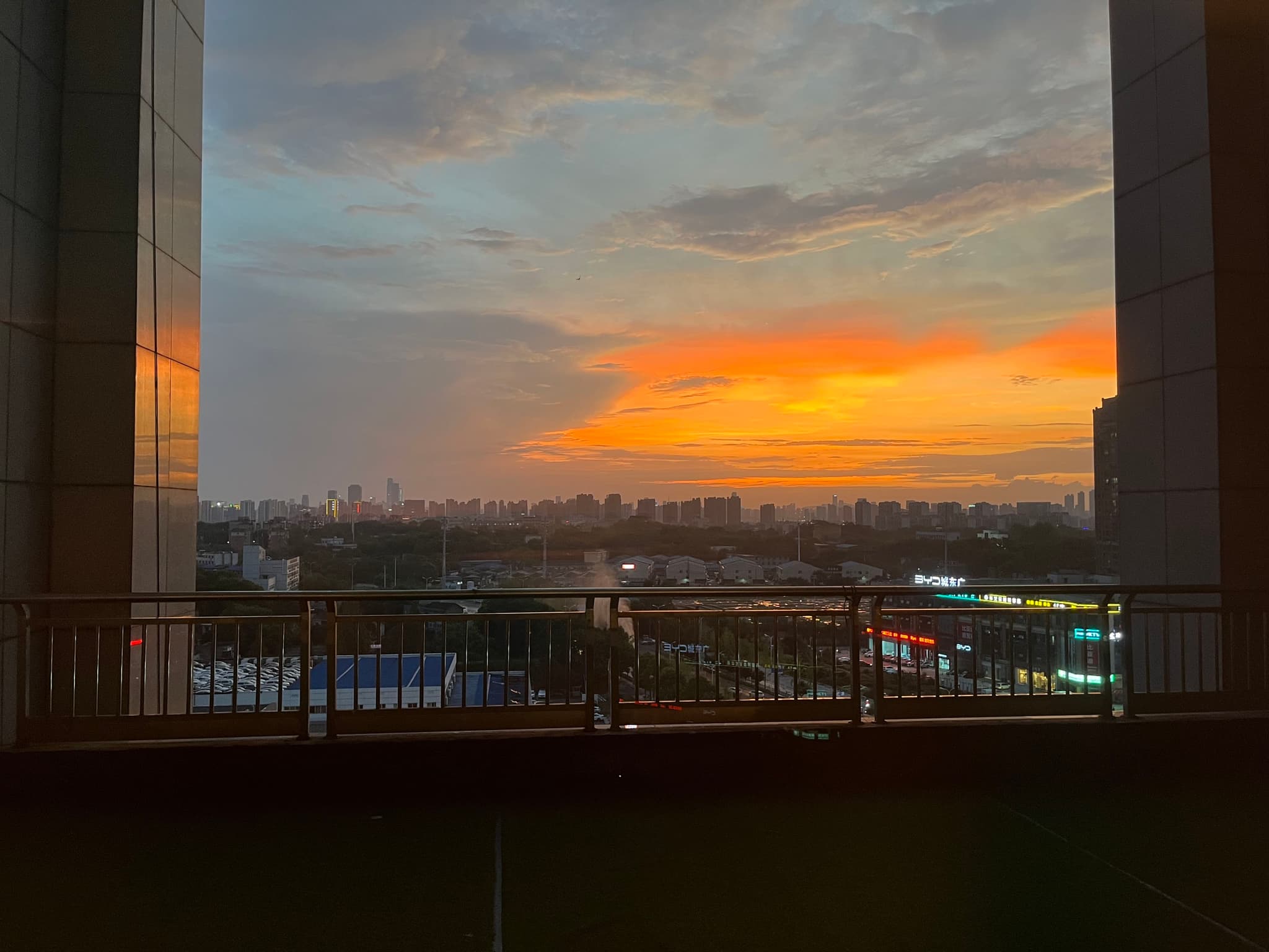 A vibrant sunset with orange and yellow hues over a cityscape, framed by two tall buildings and a railing in the foreground