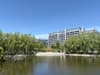 A tranquil pond surrounded by trees with modern multi-story buildings in the background under a clear blue sky