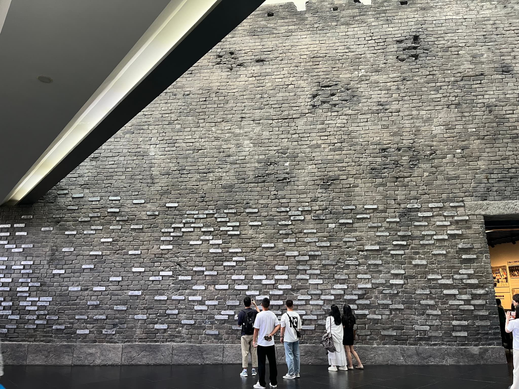 A group of people stand before a large, textured, ancient brick wall with small rectangular openings arranged in rows