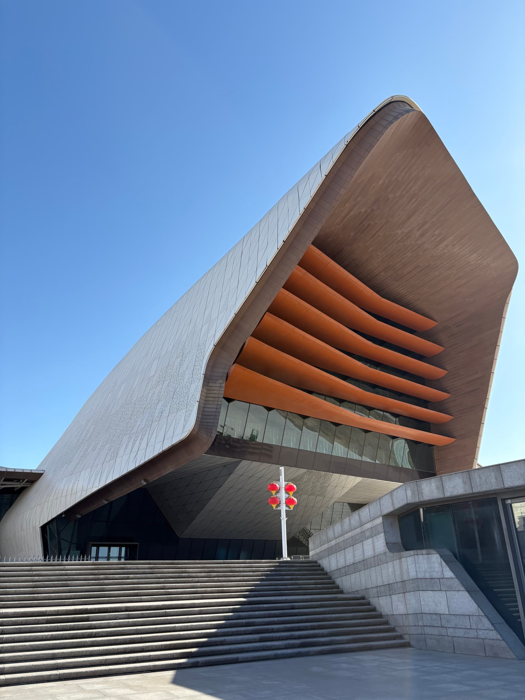 A modern architectural structure with curved lines, orange slats, and a mix of glass and concrete elements against a clear blue sky