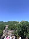 Crowds climbing a stone section of the Great Wall through forested mountain ridges beneath a blue sky