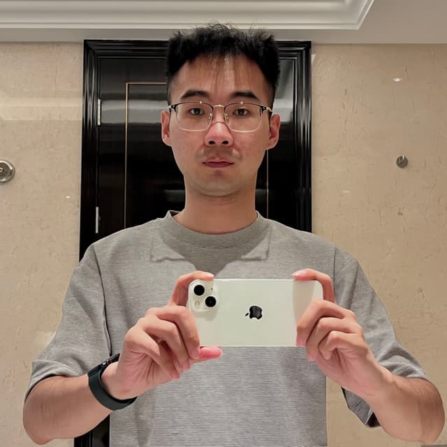 A person wearing a gray striped shirt and a black wristband holds a white smartphone, taking a mirror selfie in a bathroom with beige walls and a black-framed mirror