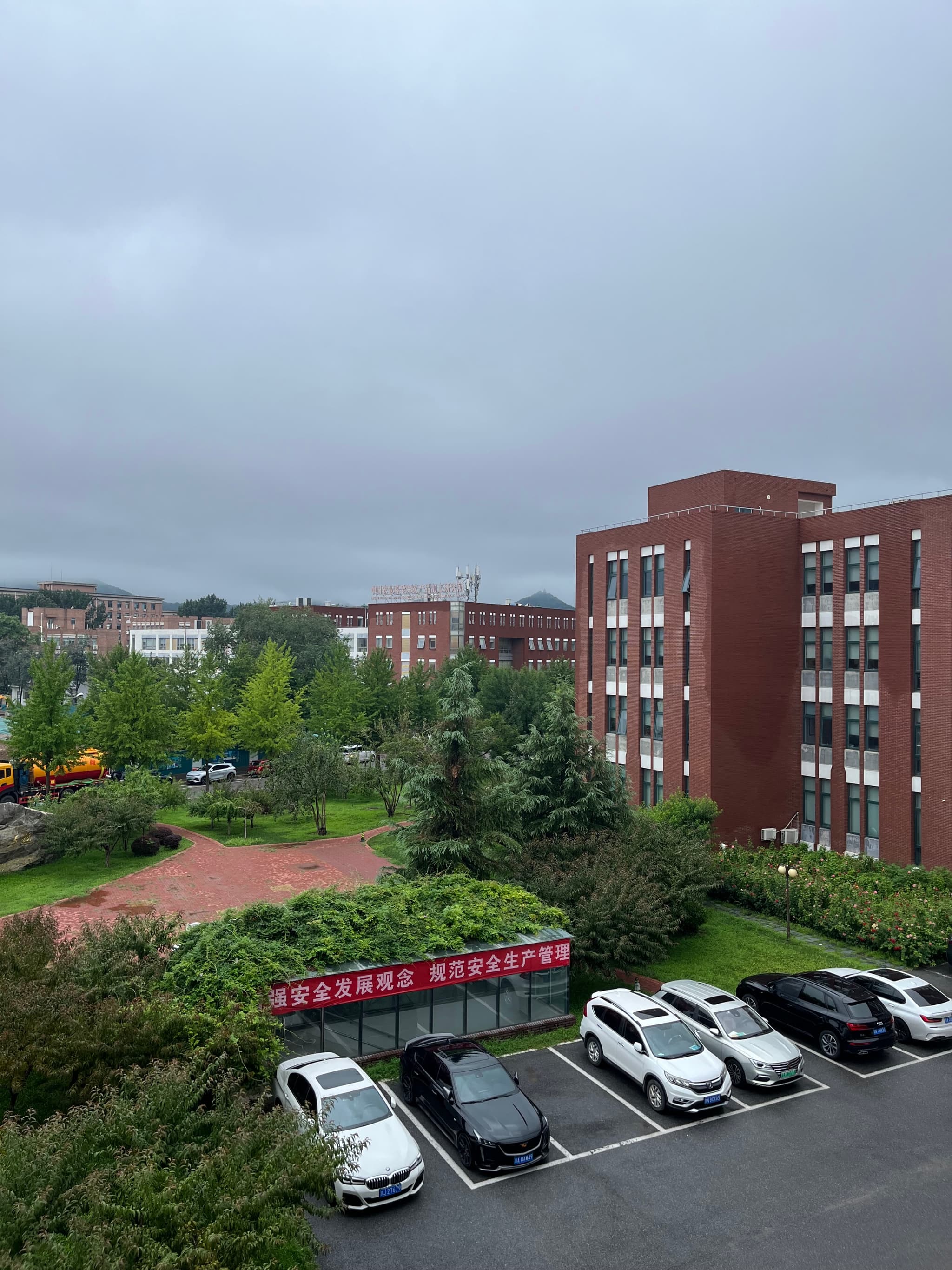 A multi-story red brick building surrounded by greenery, a parking lot with white cars, and a cloudy sky in the background