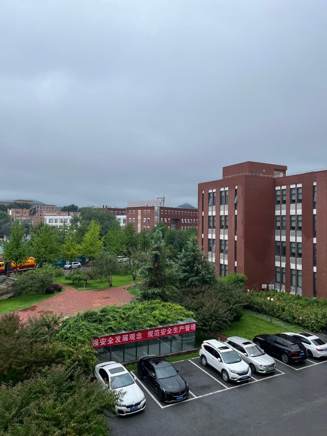 A multi-story red brick building surrounded by greenery, a parking lot with white cars, and a cloudy sky in the background