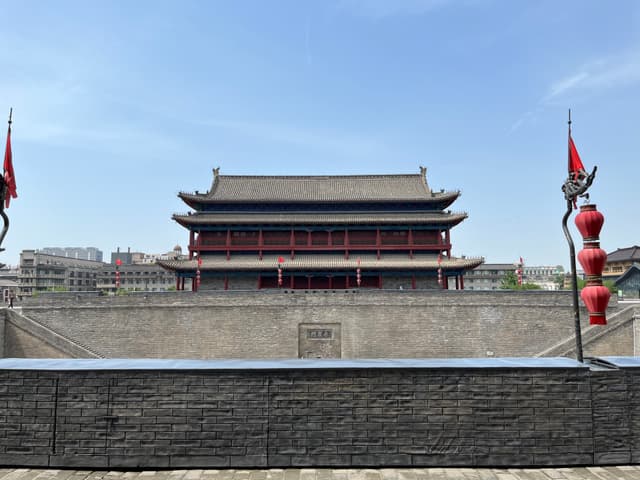 A traditional Chinese architectural structure with red accents, flanked by red lanterns and flags, set against a clear blue sky