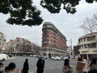 A street intersection with a distinctive triangular-shaped building, surrounded by other structures, trees, and people walking