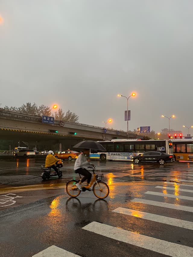 湿漉漉的街道上，行人在雨中骑行，汽车和公交车在行驶，路灯和车灯的倒影在水面上闪烁。