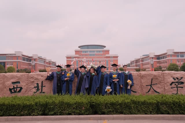 Graduates in blue gowns posing in front of a large stone with Chinese characters, with a university building in the background