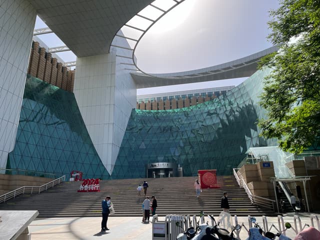 Exterior view of a contemporary building with a large, sloped green wall and a circular skylight, leading to an entrance with stairs and people