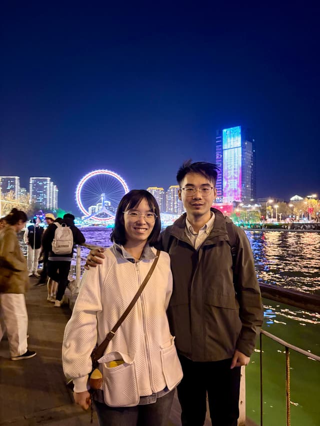 Two people standing on a waterfront promenade at night, with a brightly lit Ferris wheel and illuminated buildings reflecting on the water in the background
