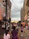 A street view of a city with buildings, pedestrians, and cars under a cloudy sky