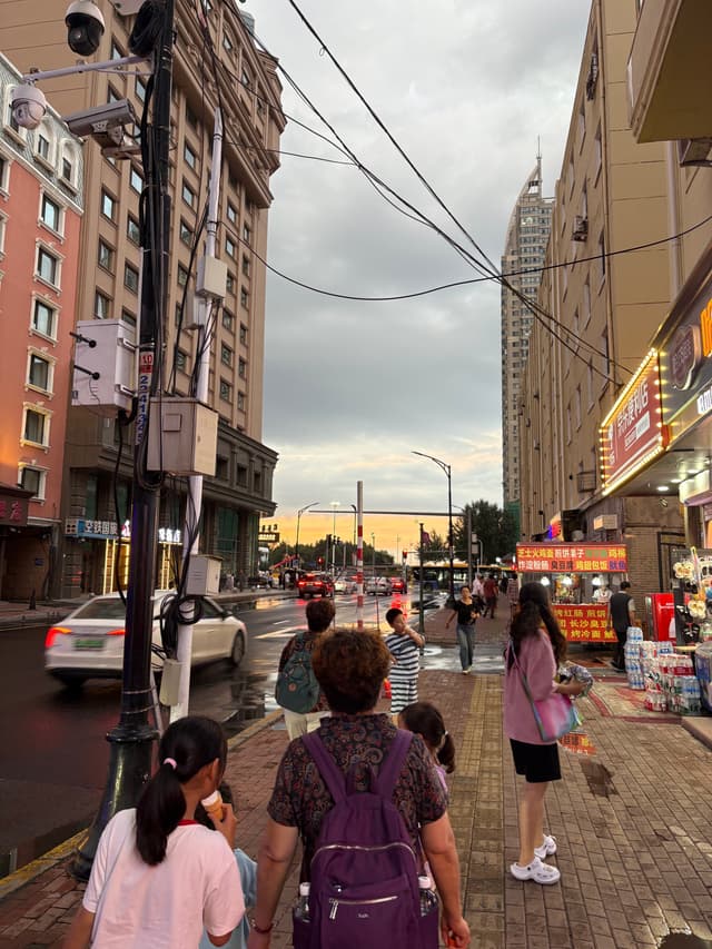 A street view of a city with buildings, pedestrians, and cars under a cloudy sky
