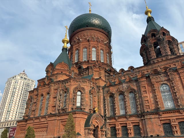A red-brick Orthodox church with green domes and golden crosses, set against a partly cloudy sky