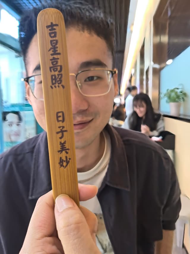 A hand holding a wooden stick with Chinese characters, indoors with a woman seated in the background near a plant