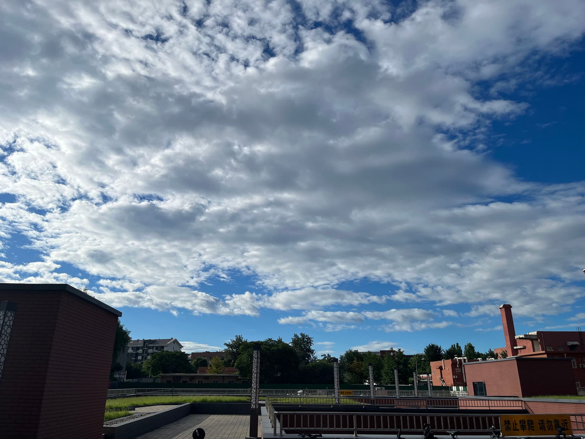 A partly cloudy sky over a suburban area with buildings, trees, and a chimney in the background