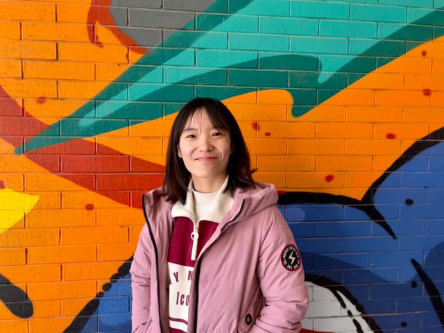 A person wearing a pink jacket and white shirt stands in front of a colorful graffiti-covered brick wall