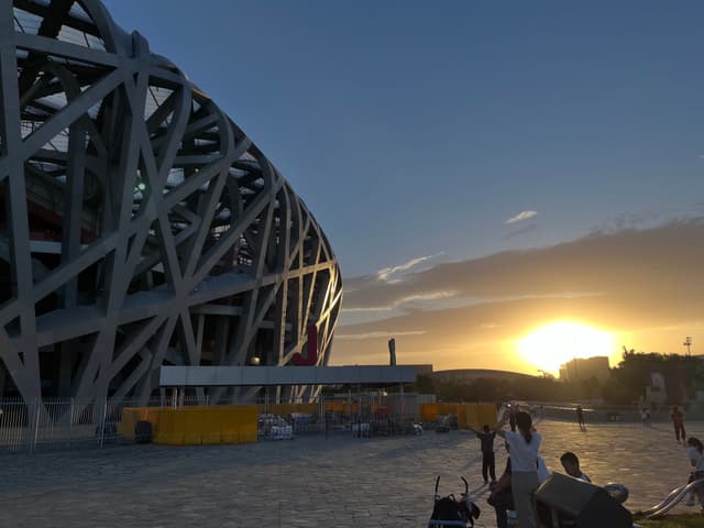 The Beijing National Stadium (Bird's Nest) at sunset, with people walking and sitting nearby