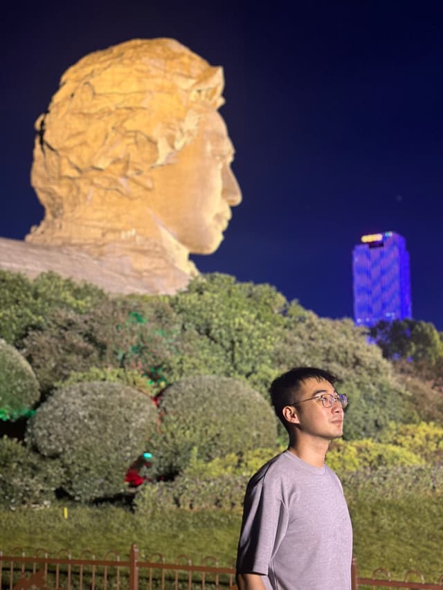 A person standing in front of a large illuminated statue and greenery, with a brightly lit building in the background at night