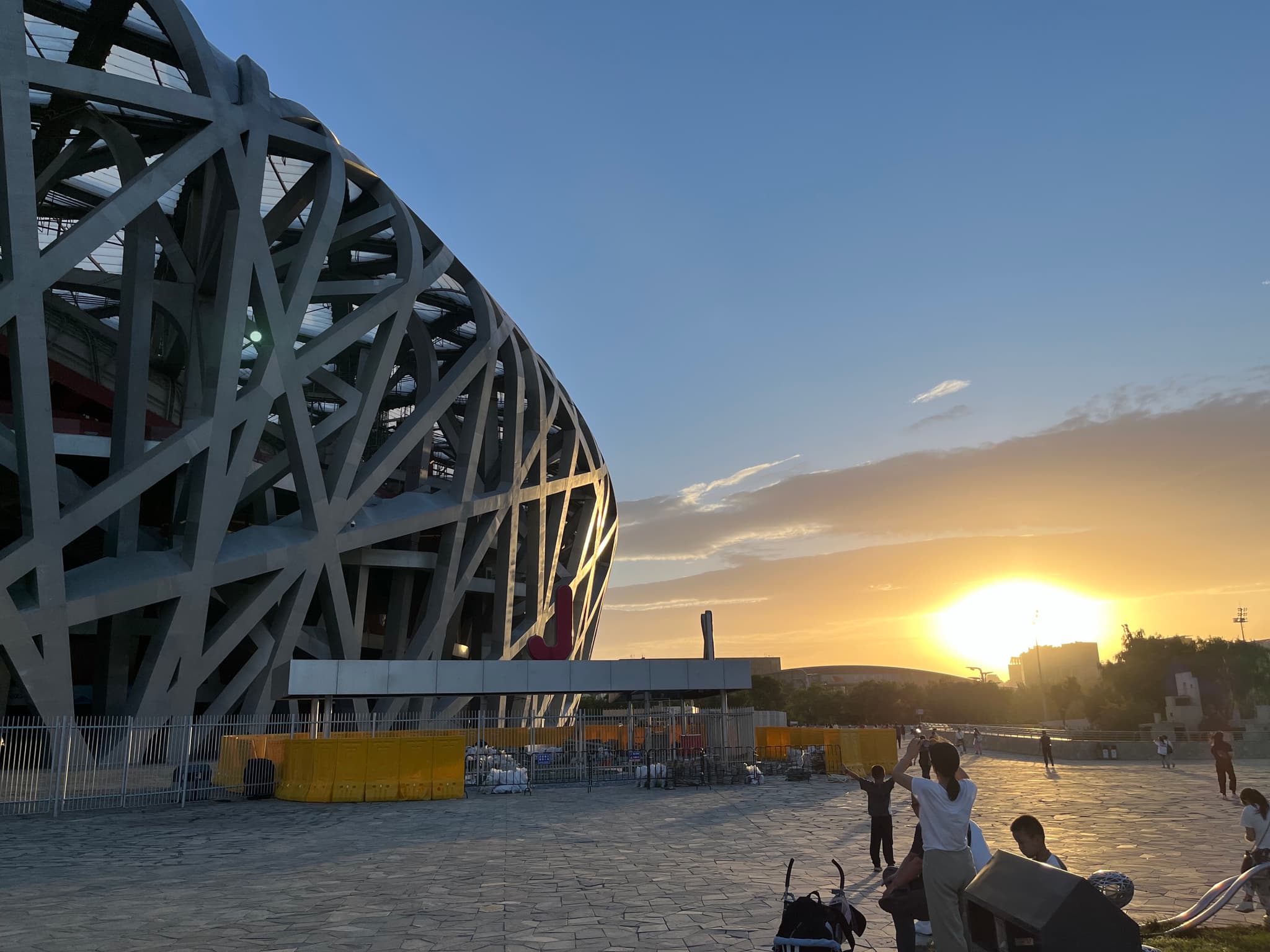 A large, modern stadium with a distinctive lattice-like exterior is shown during sunset with the sun low on the horizon