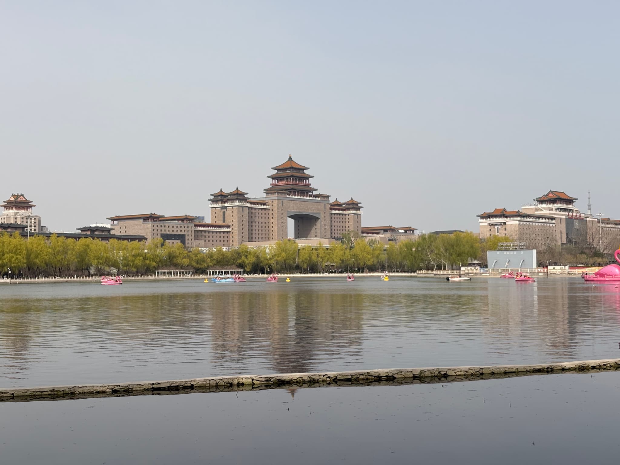 A large traditional-style Chinese building complex with pagoda-style roofs, surrounded by greenery and reflected in a calm body of water