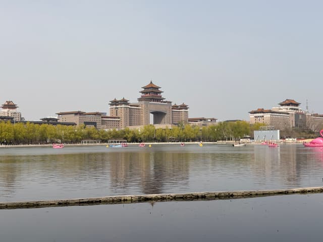 A large traditional-style Chinese building complex with pagoda-style roofs, surrounded by greenery and reflected in a calm body of water