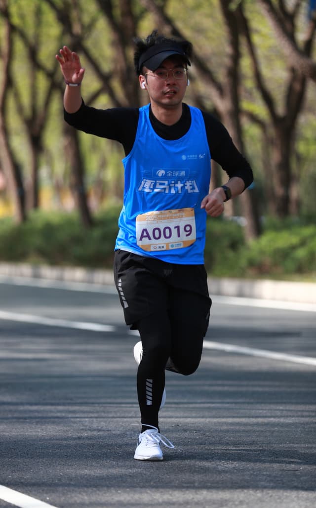 A runner wearing a blue race bib labeled A0019 and black athletic clothing is mid-stride on a paved road lined with trees