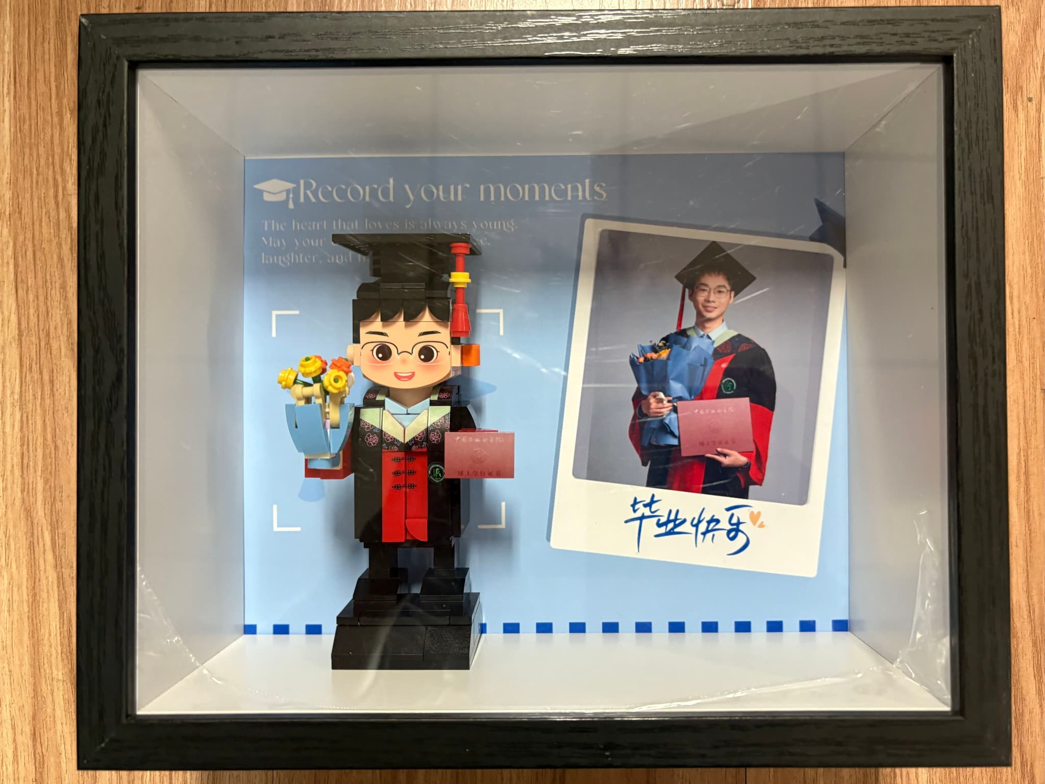 A framed display featuring a graduation-themed LEGO figure holding flowers and a diploma, alongside a Polaroid-style photo of a graduate in matching attire