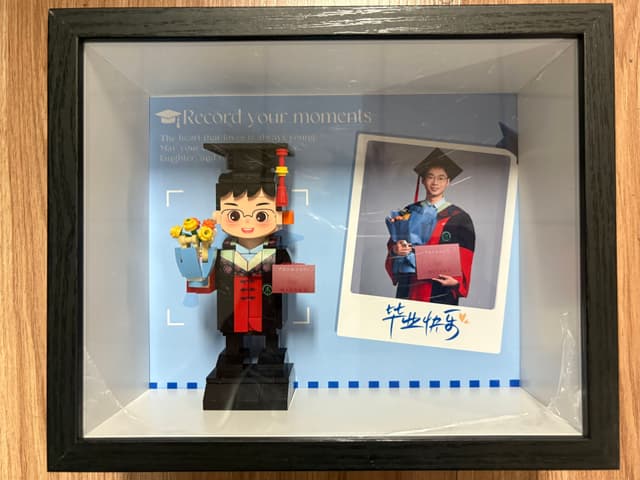 A framed display featuring a graduation-themed LEGO figure holding flowers and a diploma, alongside a Polaroid-style photo of a graduate in matching attire