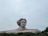 A rock formation resembling a human profile set against a cloudy sky, surrounded by sparse vegetation