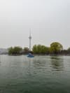 A tall broadcasting tower stands behind a body of water with a small boat on it and trees along the shore
