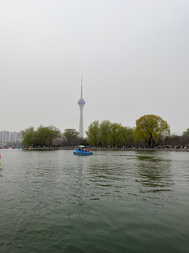 A tall broadcasting tower stands behind a body of water with a small boat on it and trees along the shore